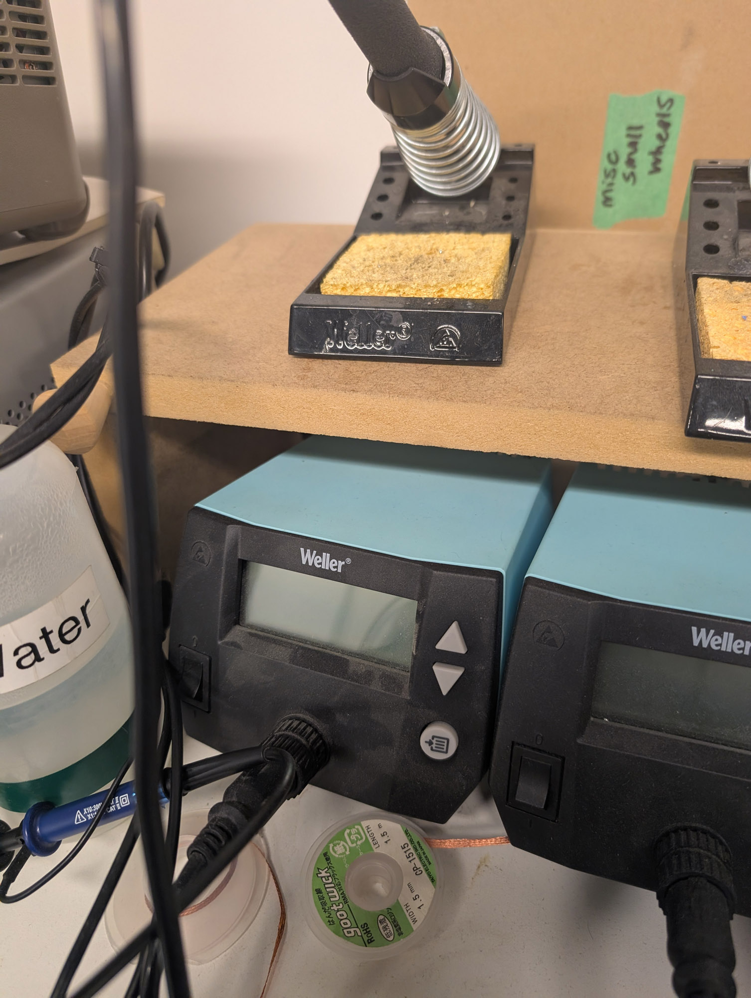 Two blue Weller soldering stations with black fronts, each with an LCD on the front and up and down arrows on each one, plus a round button with an arrow pointing to several lines. A rocker power switch is on the left, and a round handpiece connector in the middle. On a small shelf above these units is two Weller soldering iron handpieces, each in a spring-type holder with a yellow sponge